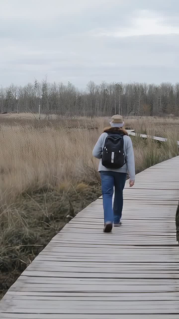 Vídeo vertical: Excursionista caminando por un paseo marítimo en dirección a los árboles con mochila negra y sombrero de ala.