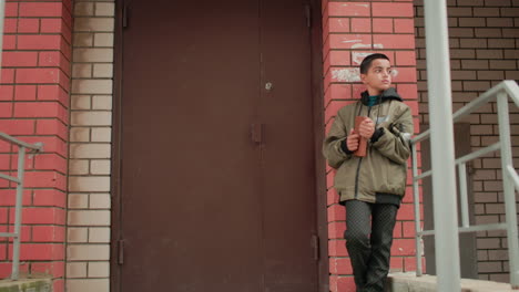 teen boy in hooded jacket leans against brick wall near building entrance holding brown book, gazing thoughtfully into distance with calm expression in quiet outdoor urban setting