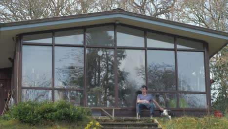 young man relaxing in front of cabin in woods with his dog