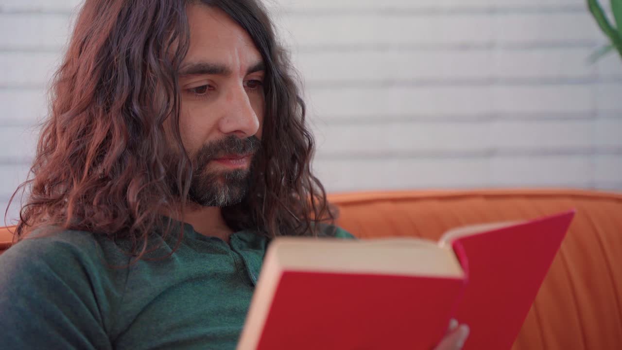 primer plano de un hombre hispano con camisa verde con cabello castaño largo y barba leyendo tranquilamente un libro rojo en un sofá naranja en una habitación blanca