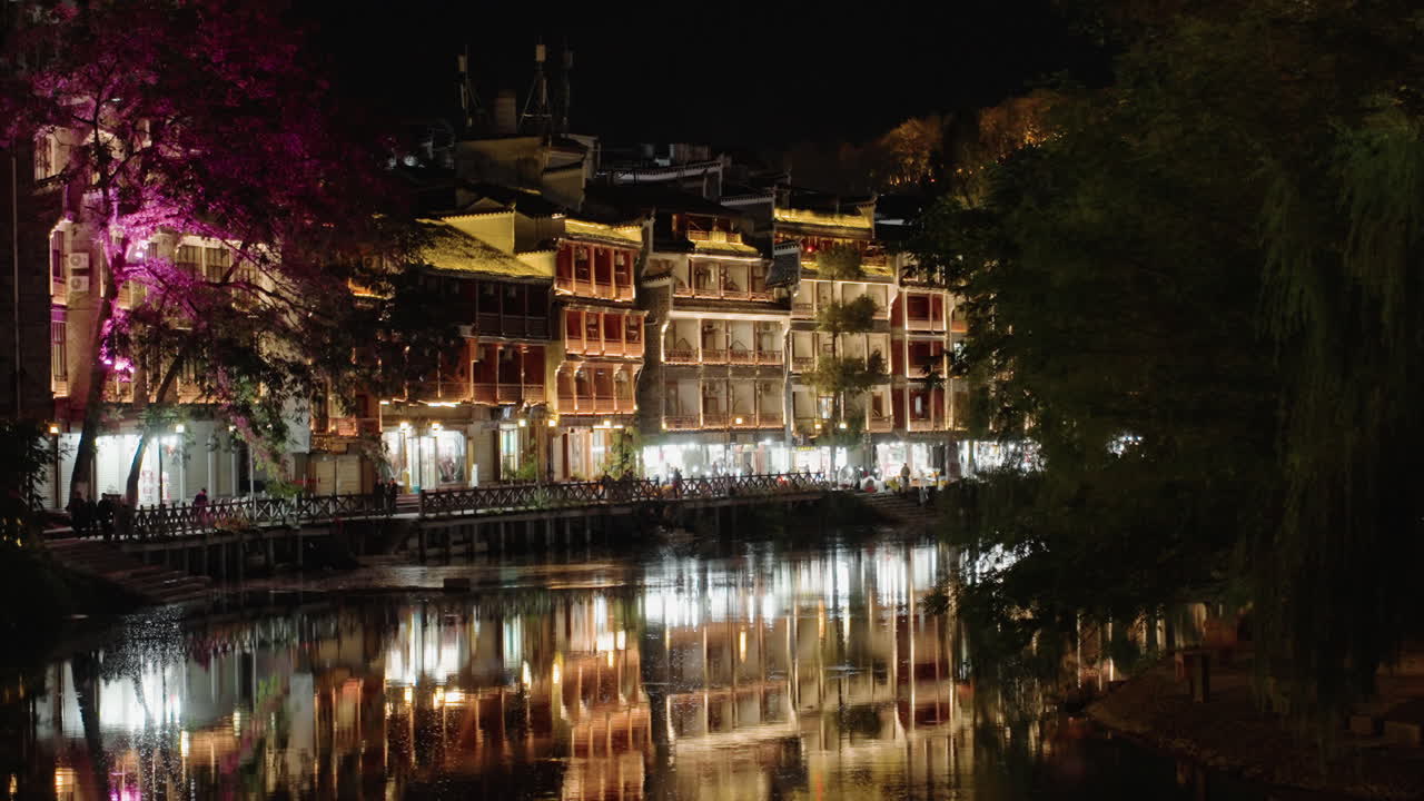 Reflexo noturno do rio com edifícios iluminados. Luzes douradas quentes projetam reflexos espelhados na água calma. Uma árvore de flores rosa brilha ao longo do calçadão, entre fachadas históricas, cafés aconchegantes e varandas.