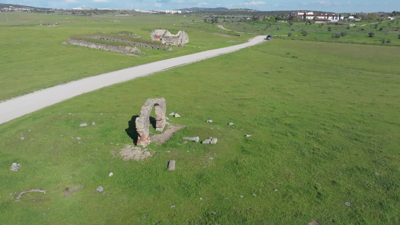 Orbital drone flight around the central arch of Puerta de Arenas. As the drone pulls back, a dirt path, remains of a pigsty, a house, and wide pastures are revealed in the rural surroundings.