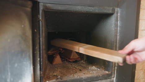 closeup of hand sliding wooden log into metal stove over glowing embers, preparing indoor fire for warm cozy winter heating, ash particles scattered on black furnace floor surface