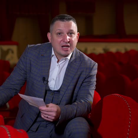 Actor at rehearsal in auditorium. Man repeating monologue while sitting among red chairs in the empty hall. Speaker rehearsing before the performance