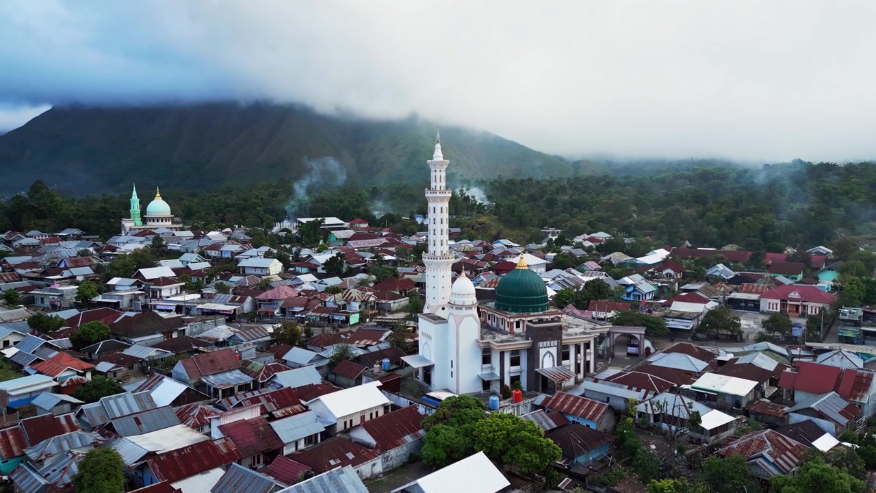 Masjid agung menjulang di atas desa pegunungan dengan asap mengepul dan puncak tertutup awan di Sembalun Lombok, rekaman drone udara di atas pedesaan Indonesia
