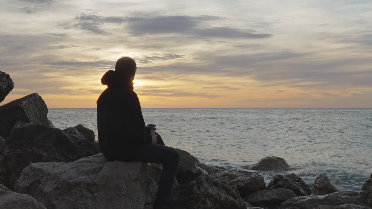el hombre entra, se sienta en una roca, abre el termo, vierte una bebida caliente y disfruta de la vista al mar durante la hora dorada