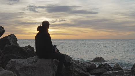 man-enters,-sits-on-a-rock,-opens-thermos,-pours-hot-drink-and-enjoys-sea-view-during-golden-hour