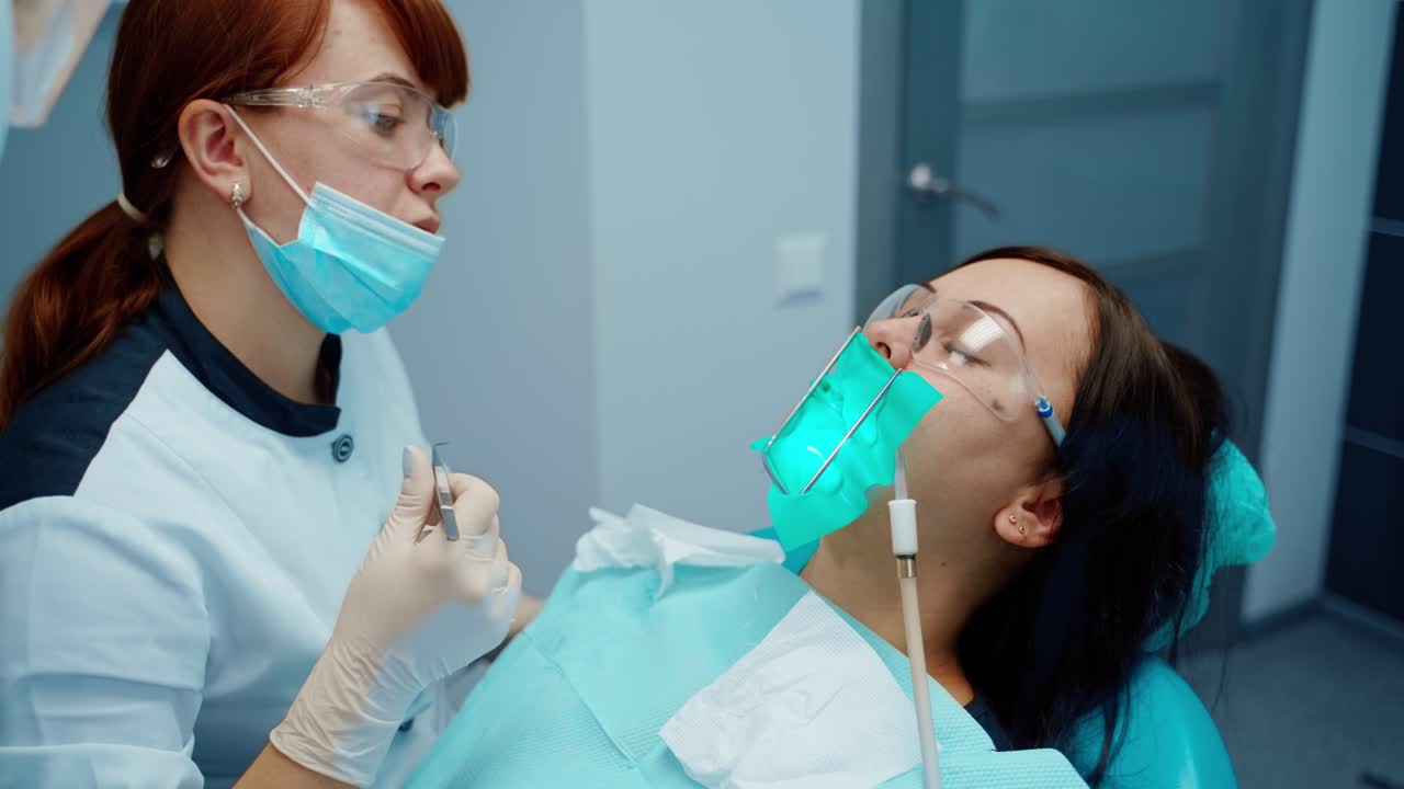 Woman patient with toothache during tooth treatment. Professional female dentist in mask and glasses treating teeth using medical instruments at stomatology clinic.