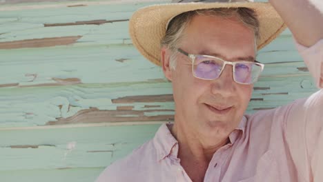 Portrait-of-happy-senior-caucasian-man-in-glasses-putting-on-hat-by-wooden-wall,-in-slow-motion