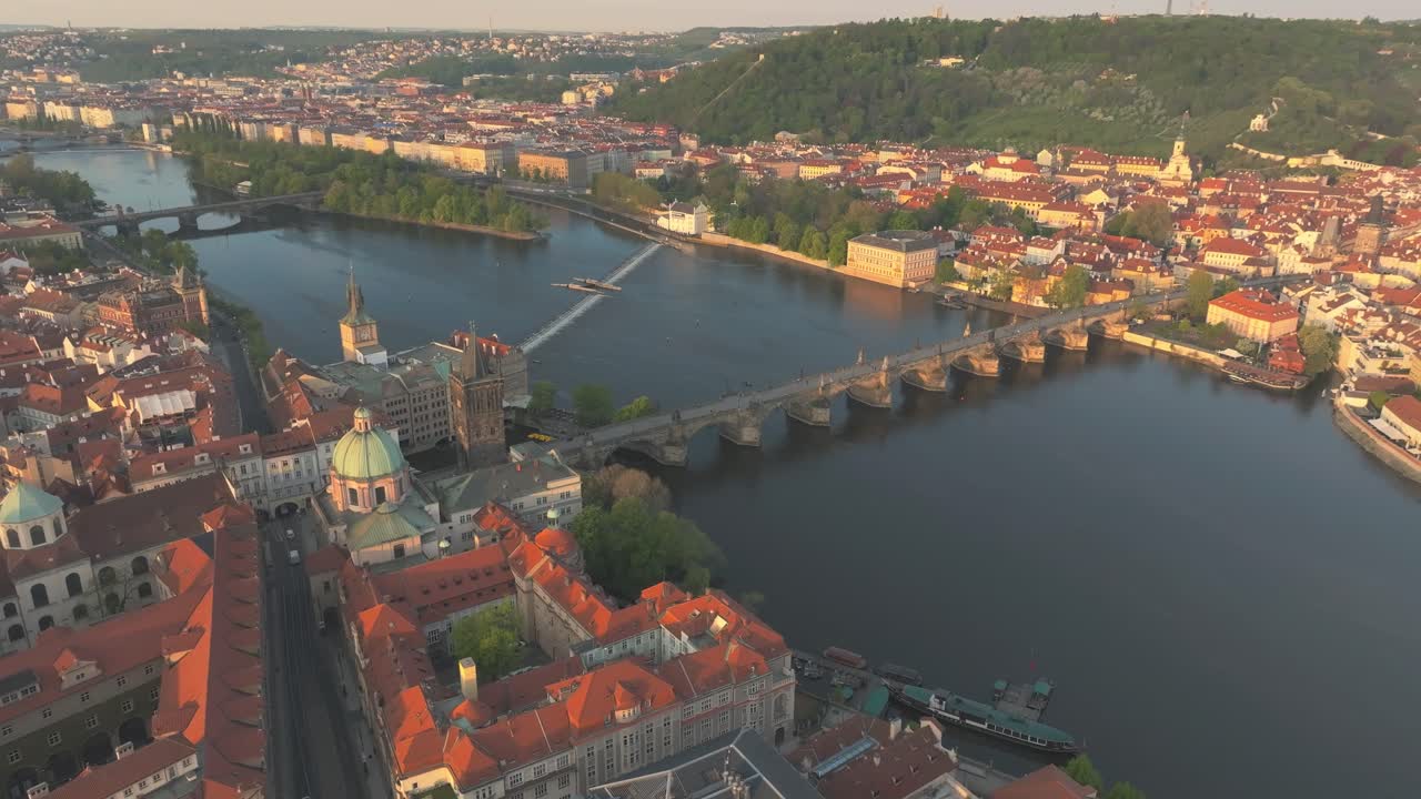 Aerial drone flyover captures a sunset cityscape of Prague, the capital of Czechia, featuring Charles Bridge spanning the Vltava River, a historic stone landmark connecting the Old and Lesser towns