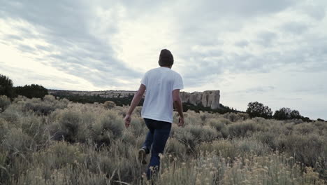 Young-man-walking-through-desert-scrub-bushes-towards-rock-formation