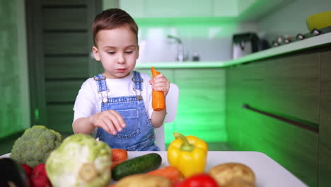 lovely four-year-old kid eating a big carrot. toddler sits at the table with numerous diverse vegetables. low angle view.
