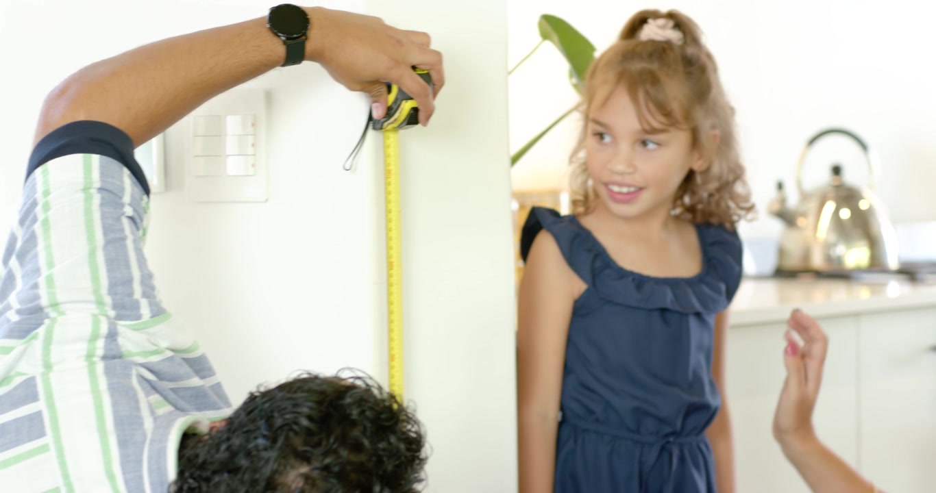Diverse family measuring daughter for growth check in kitchen, parents using yellow tape