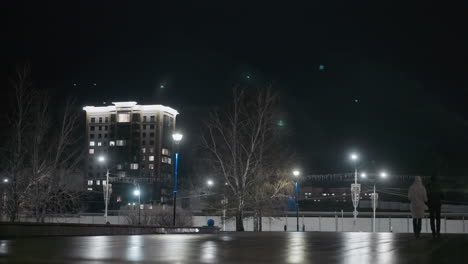 nighttime view of people walking by an illuminated urban area, with streetlights and a high-rise building in the background