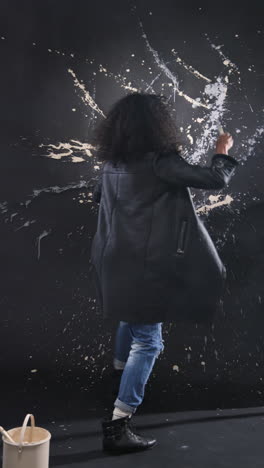 vertical video: artist creating abstract splatter painting on black wall in creative studio