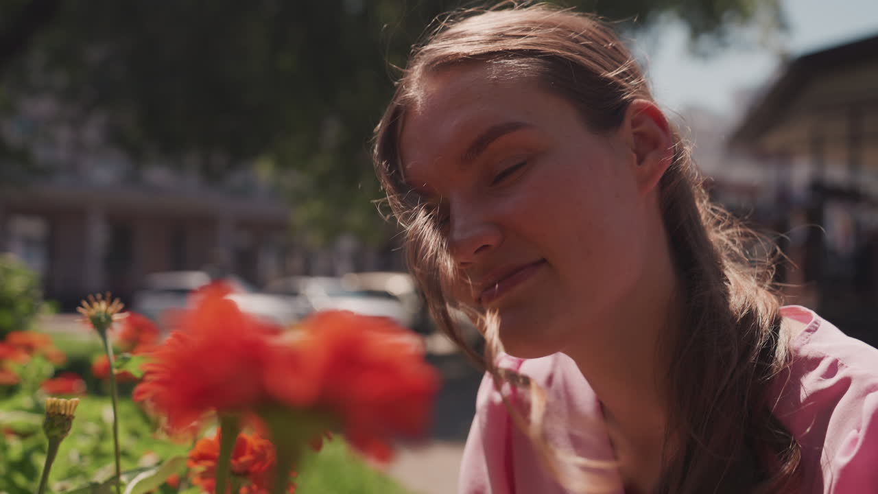 young white woman smelling red flowers, smiling in sunlit urban park, braided hair catching light, candid joyful expression, blurred storefronts and bokeh background, vibrant petals and warm afternoon