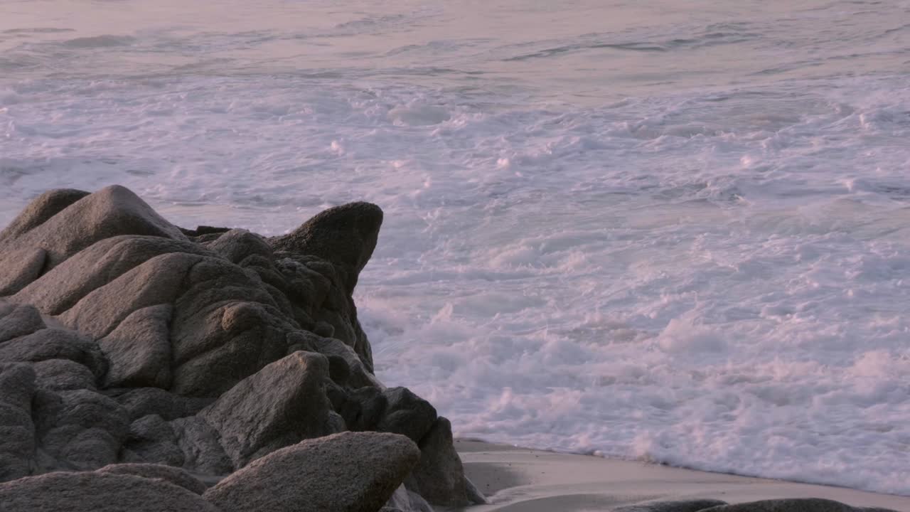 Slow-motion wide shot of waves crashing on large rocks at a sandy beach, focused on the dramatic motion of the ocean against the rugged shore.