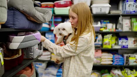 Confident-blonde-girl-with-her-small-white-dog-choosing-a-place-for-the-dog-to-sit-in-a-pet-store.-Happy-blonde-girl-in-a-light-shirt-looking-for-items-in-a-pet-store