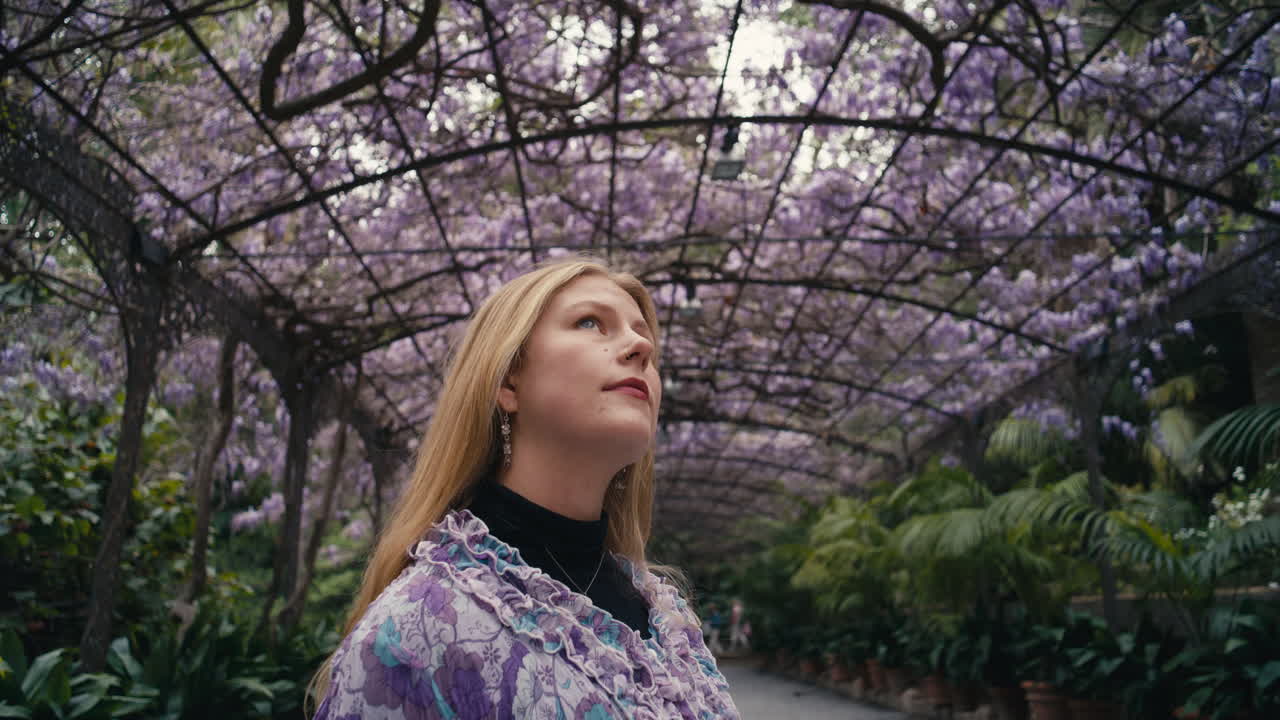 Gros plan d'une femme levant les yeux vers une glycine en fleurs aux tons violets doux et à la verdure luxuriante, créant une atmosphère calme, rêveuse et élégante