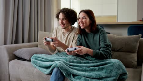 Confident-brunette-guy-with-curly-hair-plays-video-games-using-a-joystick-with-his-girlfriend-in-a-green-jacket-but-the-girl-wins-him-on-the-sofa-in-a-modern-apartment