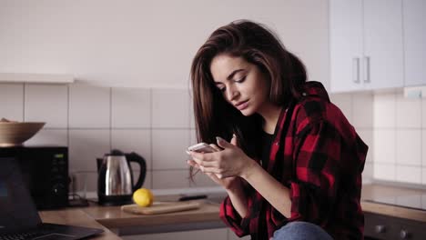 young beautiful brunette girl with messy hair in flannel shirt sitting in her kitchen and texting someone.