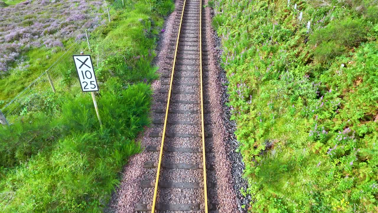 Drone camera glides above single railway track through lush Scottish Highlands, passing speed limit signs. Bright daylight, steady forward movement, vibrant green landscape