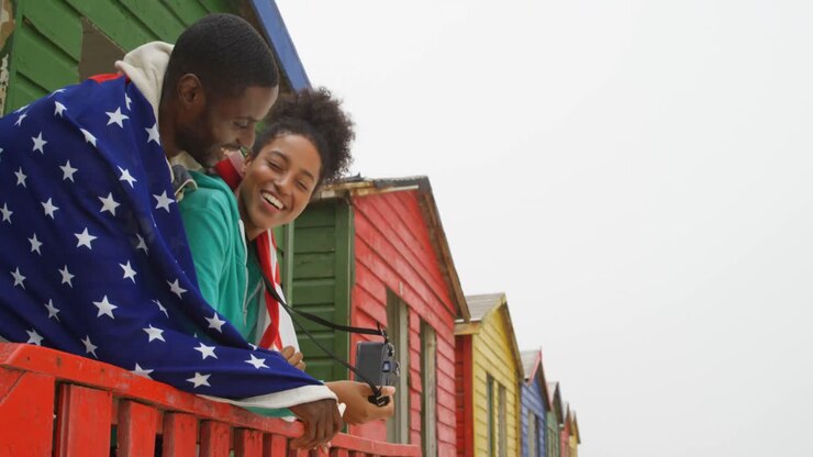 Vista laterale di una giovane coppia di colore avvolta nella bandiera americana e che si appoggia alla ringhiera presso una capanna sulla spiaggia 4k.