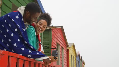 Side-view-of-young-black-couple-wrapped-in-American-flag-and-leaning-on-railing-at-beach-hut-4k