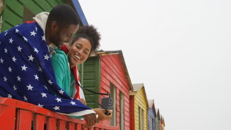 side view of young black couple wrapped in american flag and leaning on railing at beach hut 4k