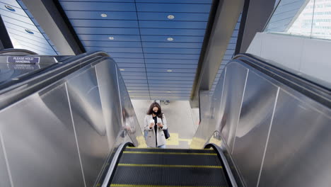 businesswoman on escalator at railway station with mobile phone wearing ppe face mask in pandemic