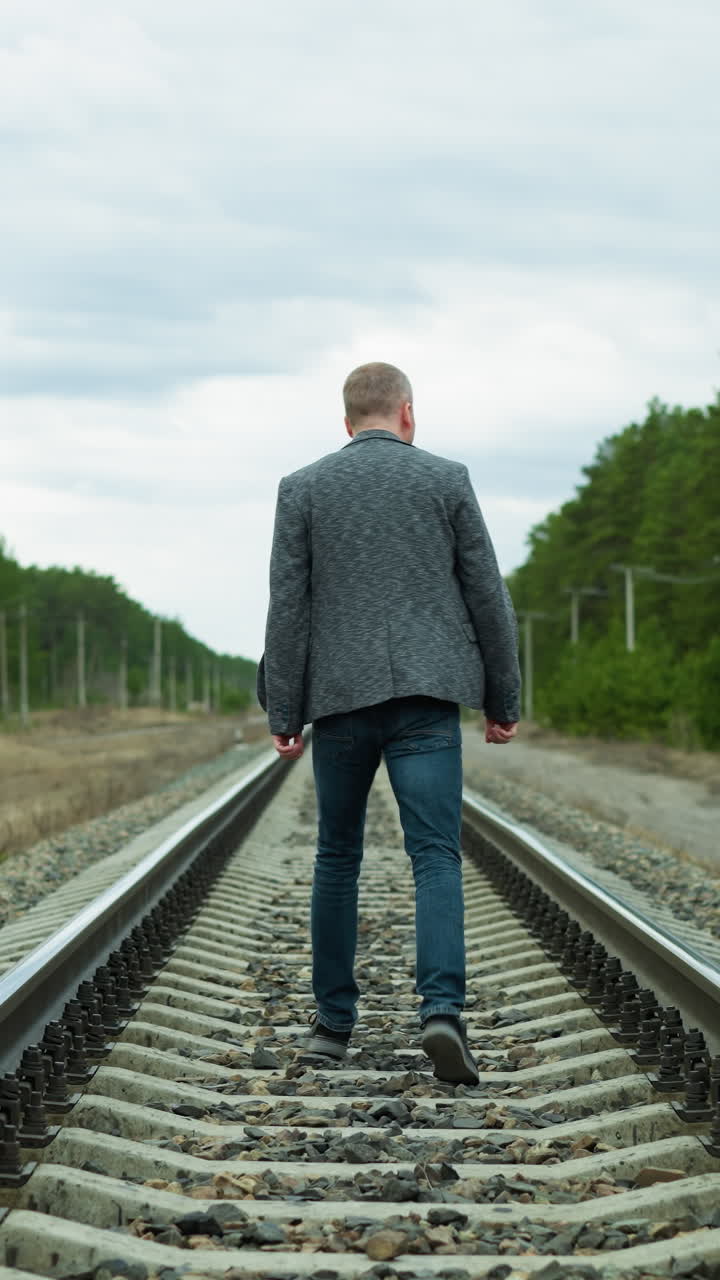 Close view of a man wearing a suit jacket and jeans, walking alone on a railway tracks surrounded by trees, with electric poles around