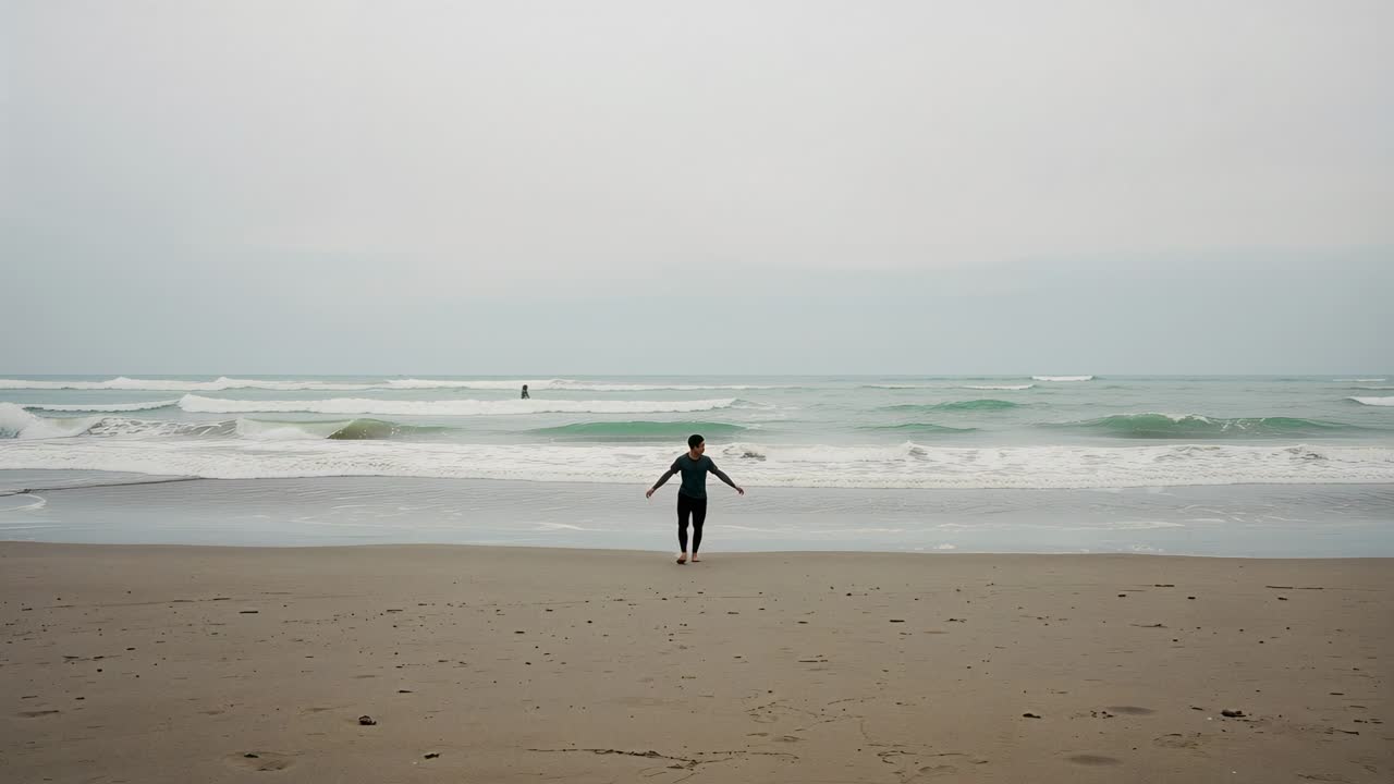 Walking man in dark wetsuit leaving breaking waves toward camera on beach to reach dry sand