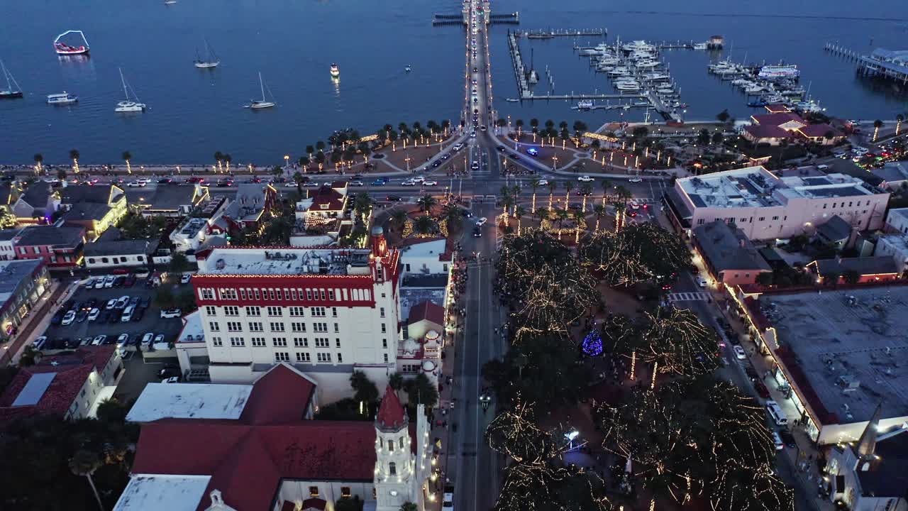 Vista aérea nocturna de una zona urbana costera, destacando un puerto deportivo con barcos, carreteras bordeadas de palmeras iluminadas y edificios cerca del frente marítimo