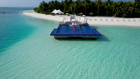 Aerial-view-of-woman-walking-in-red-swimsuit-into-infinity-pool-over-clear-blue-water-at-maldives-luxury-resort