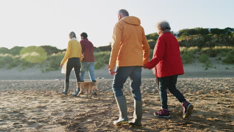 senior couple hold hands as they walk along beach with adult offspring on winter vacation