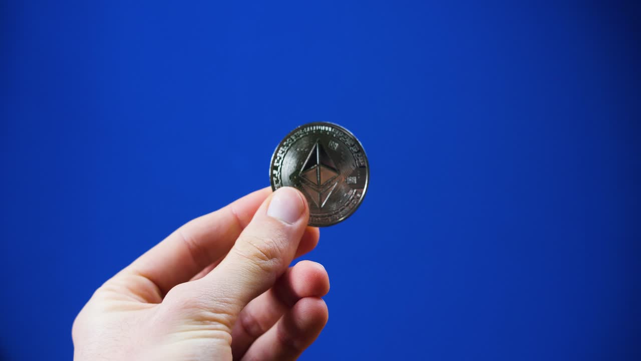 A close-up of a hand holding a silver Ethereum coin, reflecting under light with blue background