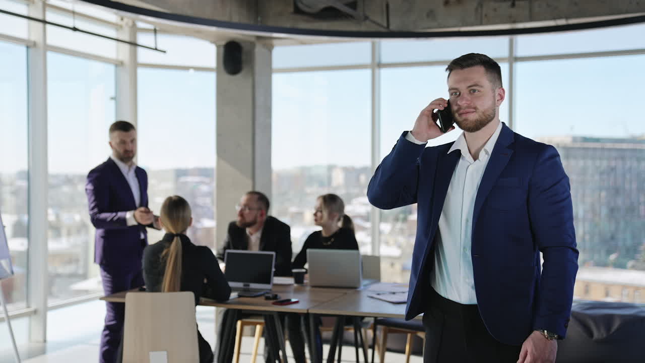 Adult businessman in suit stands at foreground and speaks on the phone. First he is calm and then laughs. Business meeting at the backdrop.