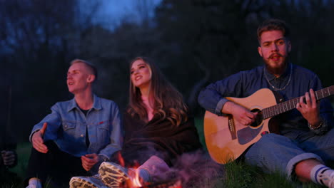 friends sit around bonfire. group of young people singing while guy playing the guitar on outdoor party in the dusk. young man playing the guitar near the fire.