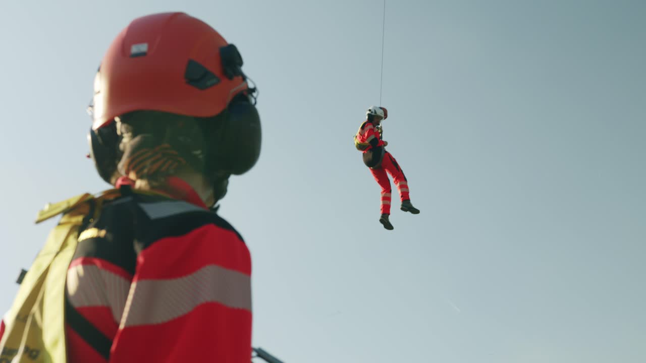 Rescuer hanging in the air from Helicopter hook and other one standing in foreground - sunny - slowmotion