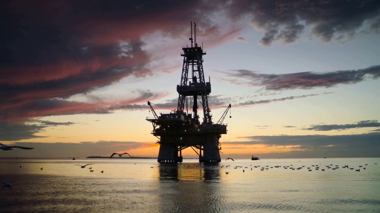 Lifting seabirds crossing sunset horizon toward offshore oil rig, service vessel and calm ripples