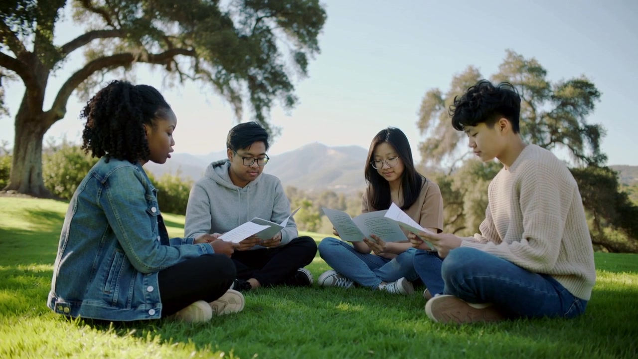 Opening booklets, four students turning pages and sharing glances in park, for study, denim jacket