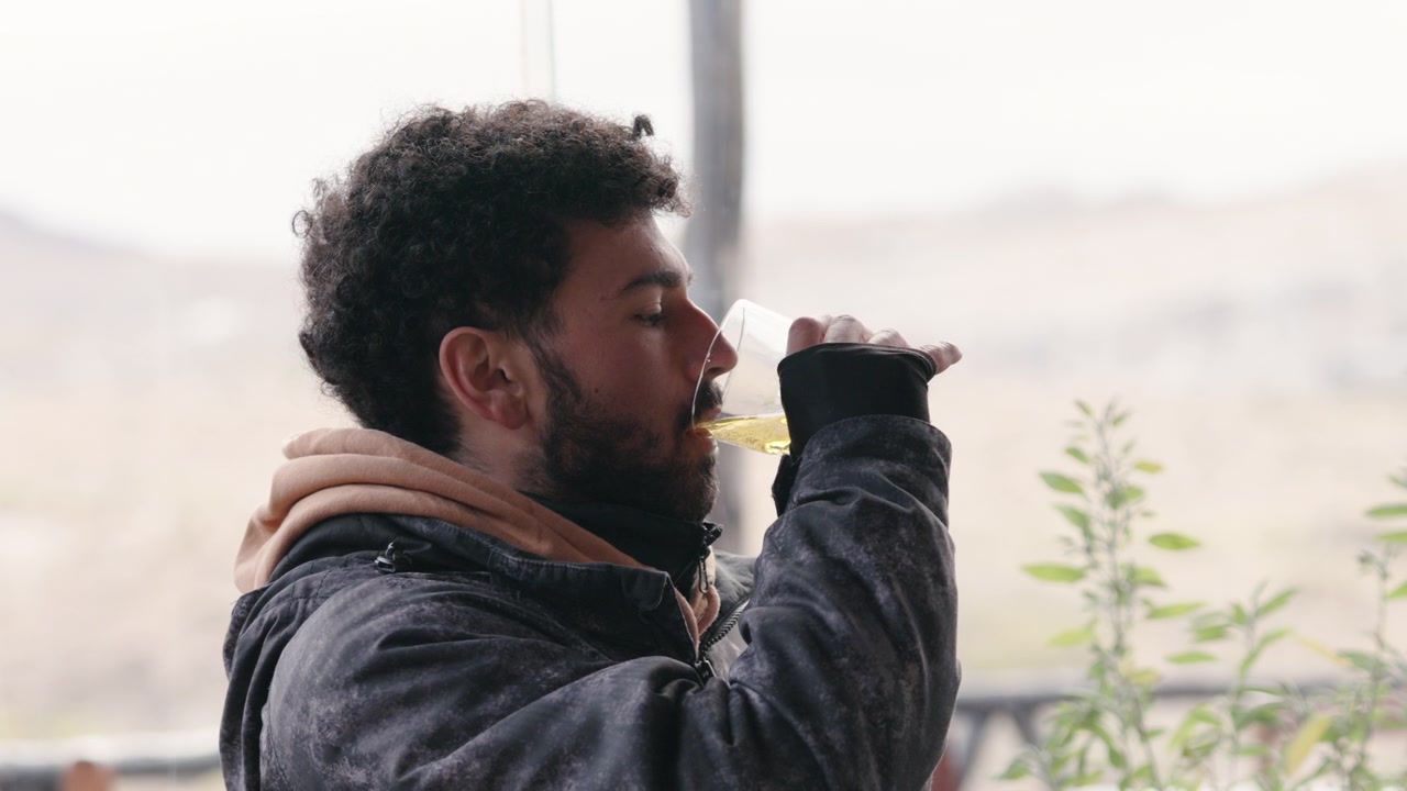 A man in a warm jacket sips a drink while seated at a restaurant with a mountain backdrop. Ideal for use in lifestyle or travel content.