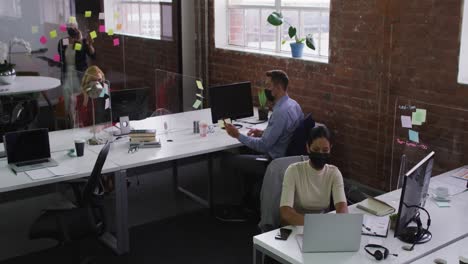 Diverse-male-and-female-business-colleagues-wearing-face-masks-using-laptop-talking-on-smartphone