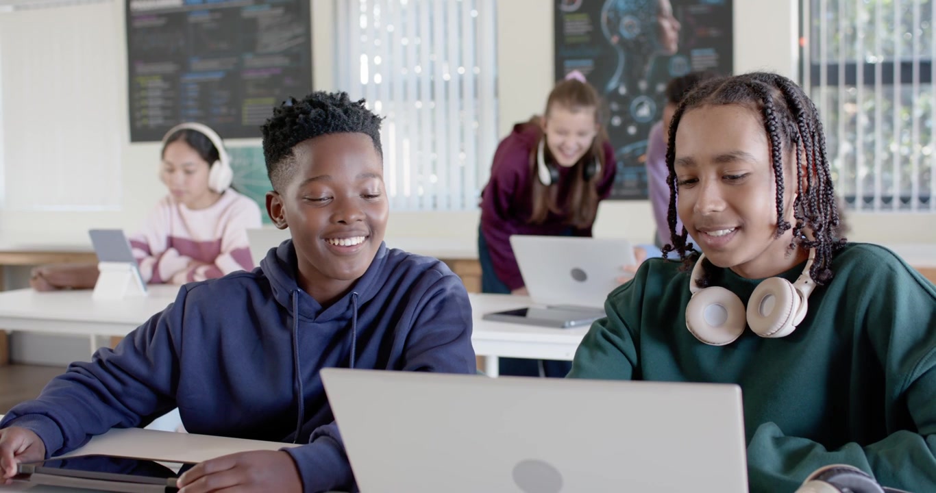 Two teenage boys exchanging glances, working on laptop in classroom, right teen pointing and typing