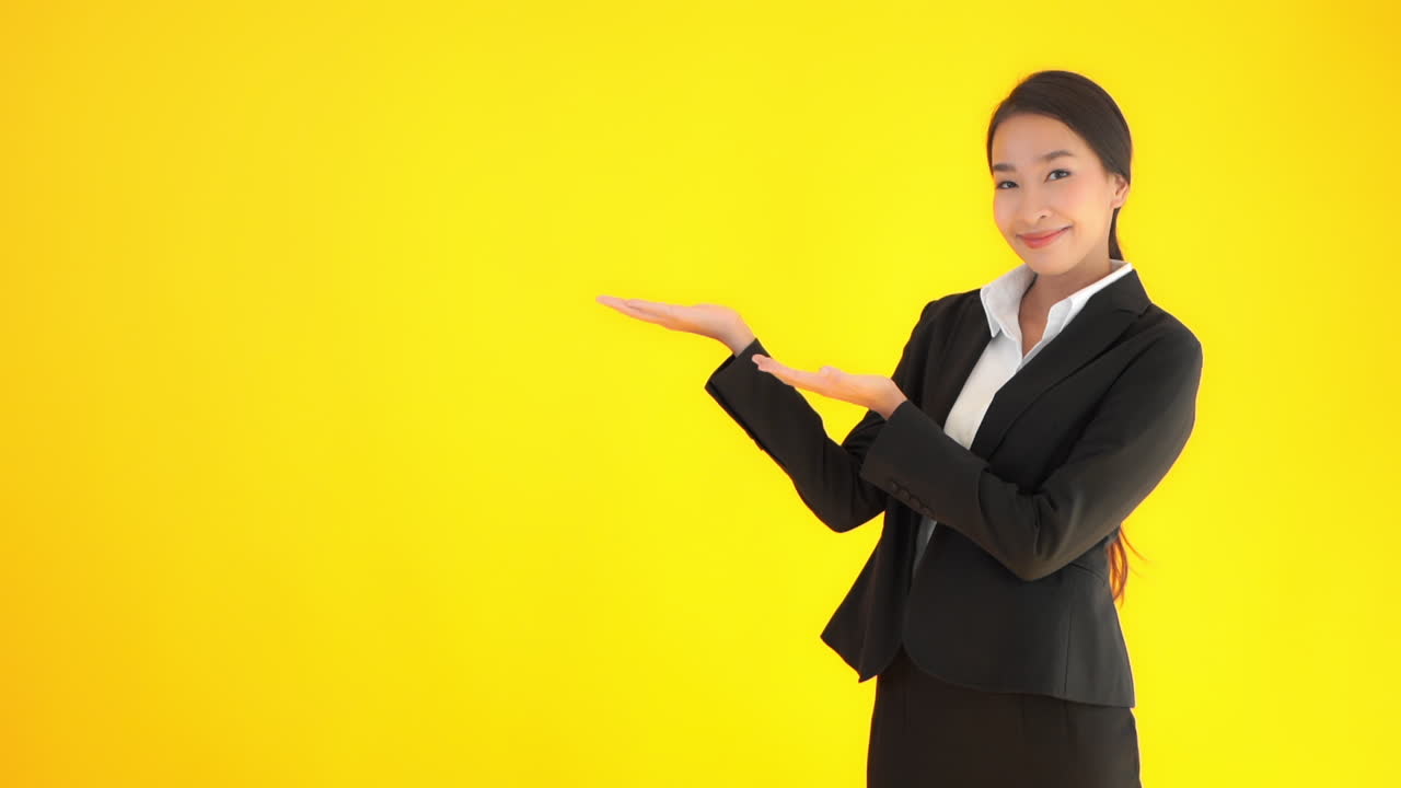 A young, attractive, Asian woman standing against a yellow wall and dressed in a business suit gestures with her hands as if pointing out a product. Copy space