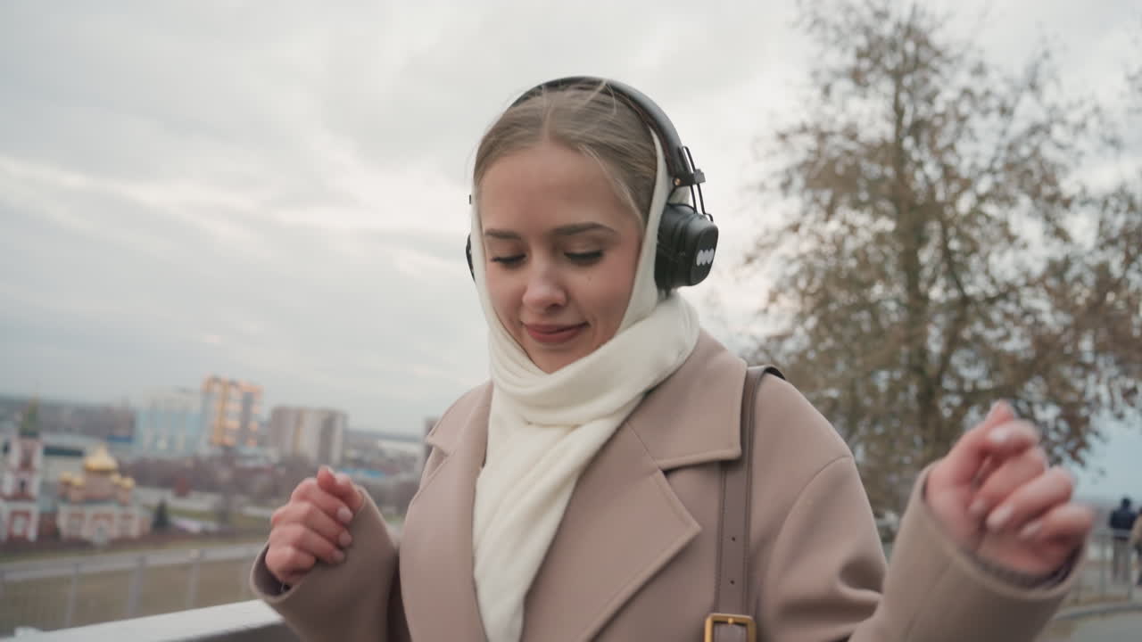 Danza gioiosa al fiume. Donna balla in riva al fiume con le cuffie. Donna aggraziata si muove lungo la ringhiera del fiume a ritmo di musica. Donna in cappotto beige esprime gioia ballando vicino all'acqua con le cuffie.