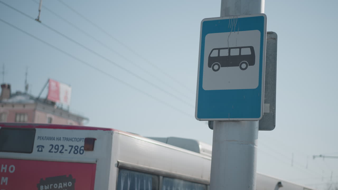 close view shows bus stop pole while bus passes along city street, overhead wires cut blue winter sky, blurred buildings behind, urban transit moment with motion, symbol foreground for commute scenes