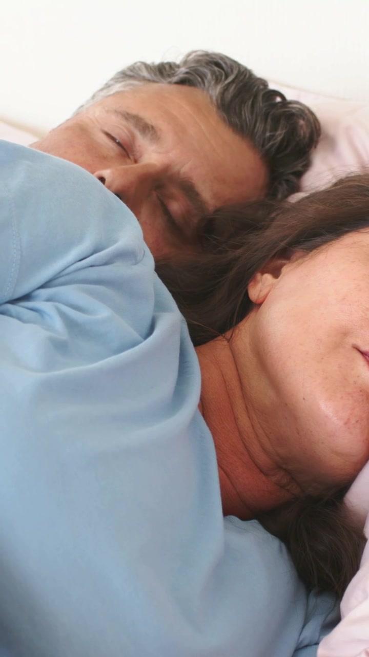 Vertical video: Camera starting filming older couple breathing while resting on bed with blue tops