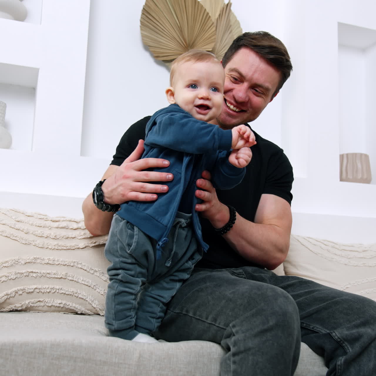 Uomo caucasico felice seduto sul divano tiene in braccio un bambino. Un bambino carino e sorridente sta in piedi sostenuto dal padre. Vista dal basso.