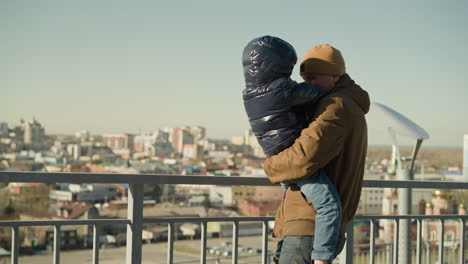 a father tenderly holds his son close to an iron railing, both smiling looking at each other warmly, with cityscape in view, with both dressed warmly in jackets